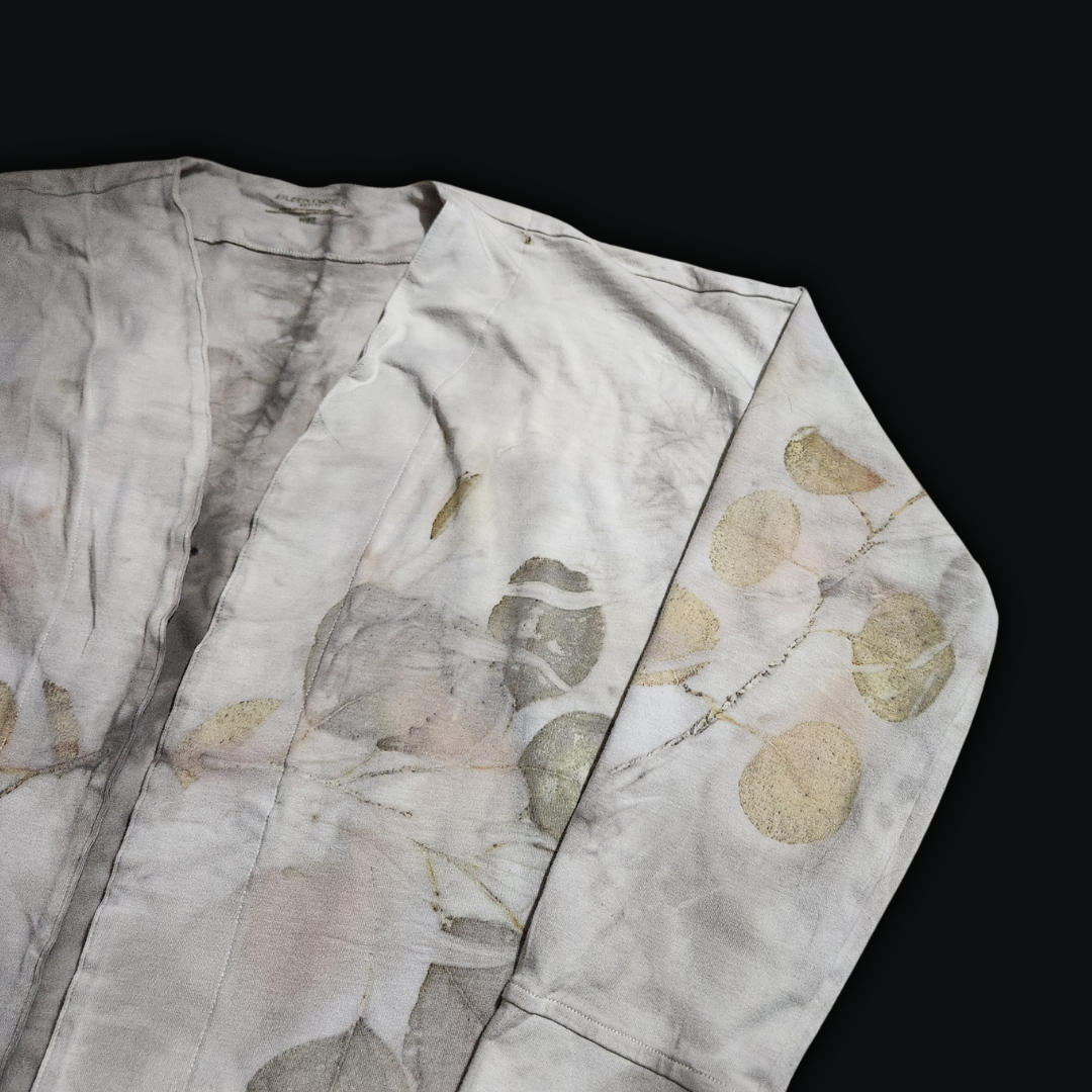 A delicate, light-colored long-sleeved cardigan, its fabric kissed with a soft, earthy pattern of eucalyptus leaves. The cardigan lies flat against a dark backdrop, reminiscent of the shadows beneath a canopy of trees.