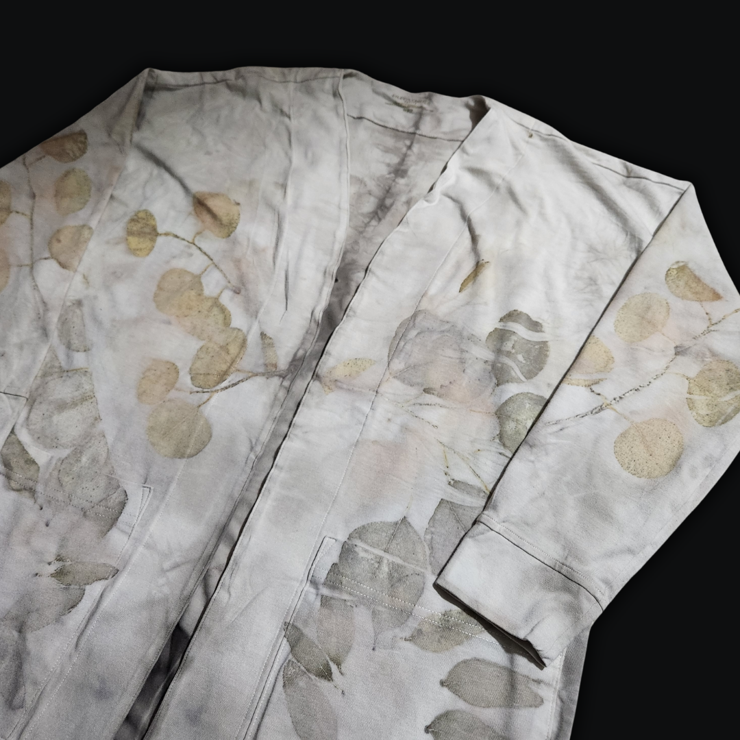 A light-colored long-sleeved cardigan, its fabric delicately patterned with eucalyptus leaves, a testament to the beauty of nature's artistry. The cardigan rests against a dark background, capturing the quiet elegance of a forest clearing.