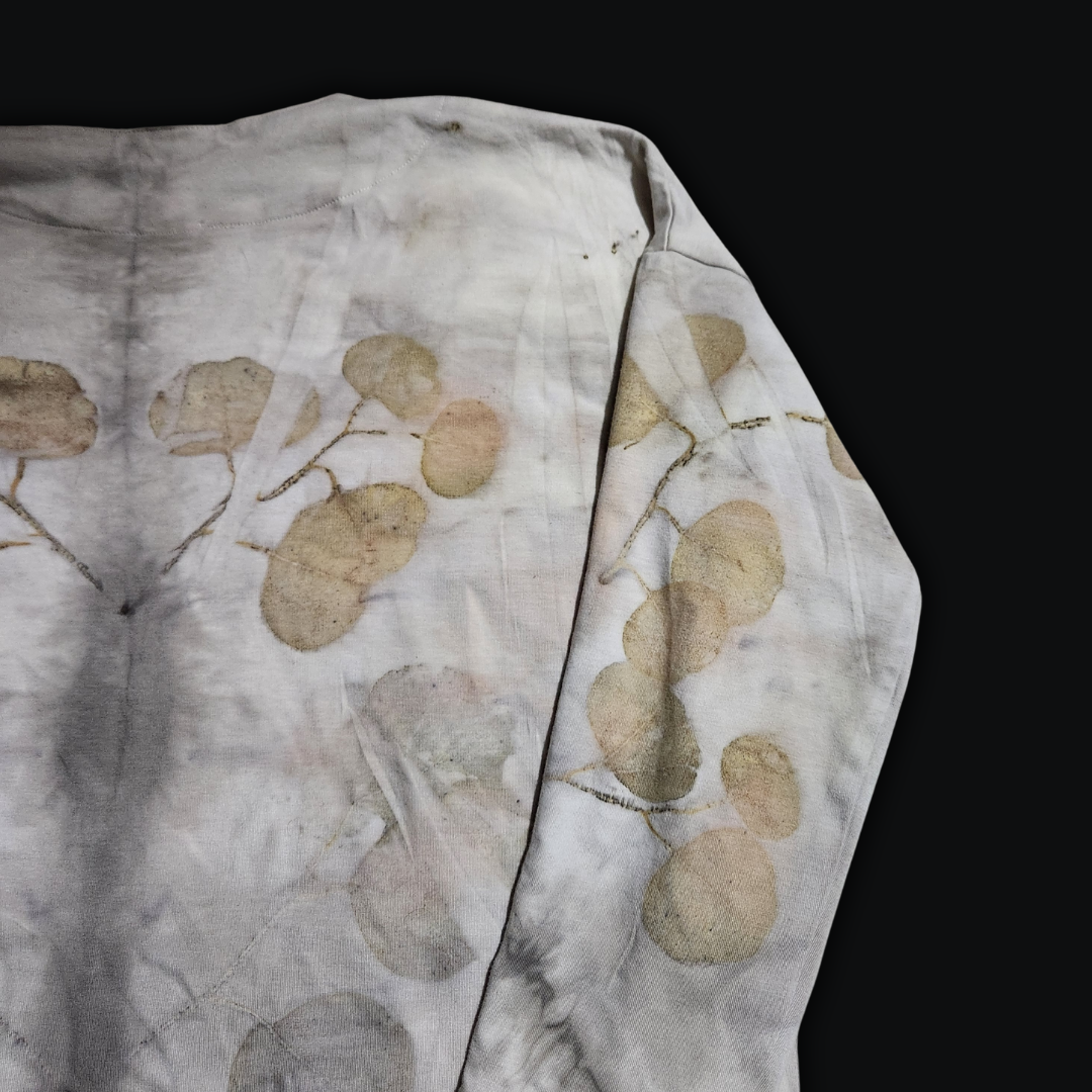 A light-colored long-sleeved cardigan, its back adorned with a subtle, leafy pattern, a whisper of the forest woven into the fabric. The shirt lies flat against a dark backdrop, inviting the viewer to imagine the gentle rustling of leaves in a quiet woodland.